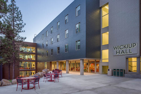 Exterior of Wickiup Hall at dusk, a four-story residence hall with warmly lit windows and a patio with red tables in the foreground