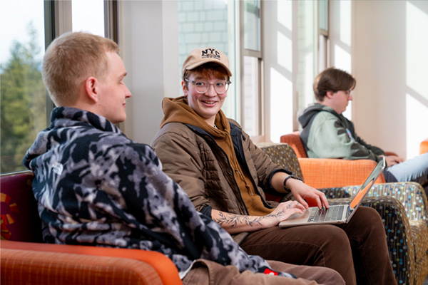 Three students relax with laptops in a bright lounge, two sitting together and talking while a third works nearby.