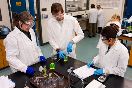 Group of students conducting a chemistry lab experiment, measuring and recording results while wearing lab coats, gloves, and safety goggles