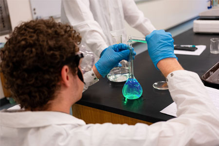 Student conducting a chemistry experiment, pouring green liquid into a flask while wearing gloves and safety goggles