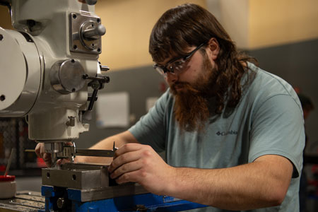 A student wearing safety glasses carefully operating a milling machine, adjusting a metal workpiece in a machining lab.