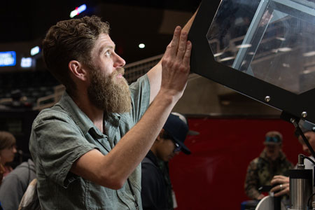 A manufacturing technology instructor examining a machine component up close, lifting a protective cover while students observe in the background in a workshop setting.