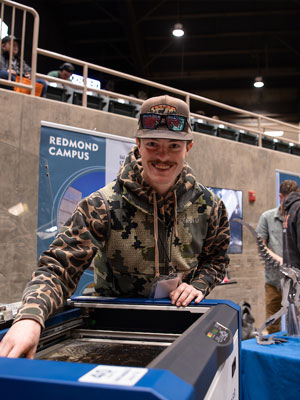 A student smiling while operating a machine at an indoor campus event booth, with a “Redmond Campus” sign visible in the background.