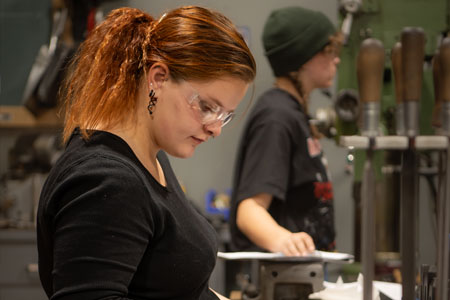 A student wearing safety glasses concentrating on a metal project while another student works in the background of a workshop.