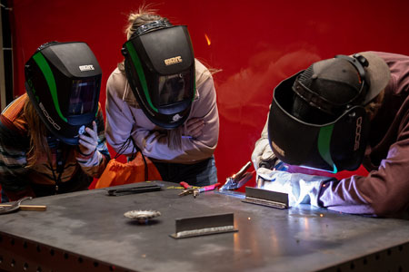 Three students wearing welding helmets working together at a metal table, actively welding and observing the process.