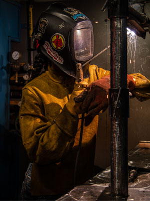 A welder wearing a protective helmet and gloves working on a vertical metal pipe, with bright sparks visible during the welding process.