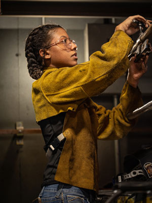 A student wearing a welding jacket and safety glasses adjusting a metal component in a workshop environment.