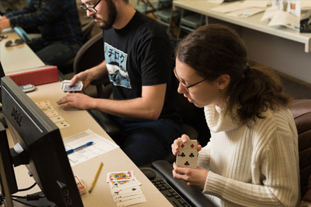 Two students working with playing cards at classroom desks during a math lesson, with computers and papers nearby.