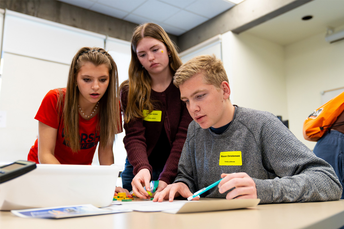 Three high school students lean over a table, focused on solving a math problem using small colorful blocks and a worksheet during the COCC Math Contest.